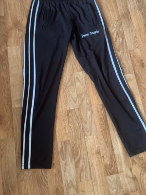 Palm Angels svarta mjukisbyxor - Palm Angels svarta mjukisbyxor med vita dubbla sidoremsor och broderad logga på vänster sida. Byxorna har elastisk midja och raka ben. Perfekta för en avslappnad och trendig streetwear-look.