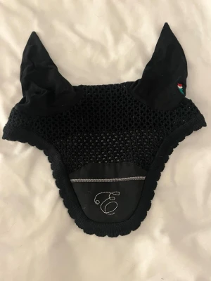 Svart huva till häst Equiline  - Säljer en svart huva till häst med nätad mittdel och tyg över öronen. På ena örat finns en liten broderad detalj i rött, vitt och grönt. Framtill finns en elegant brodyr med bokstaven E och en tunn silverrand.
