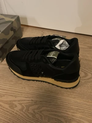 Valentino rockrunners - Stilrena svarta rockrunners från Valentino med mattsvarta nitar på sidan och beige sula. Skorna har klassisk låg profil, snörning och detaljer i både läder och textil. Perfekta för dig som gillar exklusiv streetstyle.