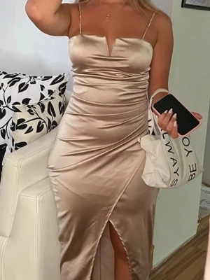 Satin BALKLÄNNING /bröllopsklänning - Snygg långklänning i champagnefärgad satin med smala axelband och en unik v-ringning. Klänningen har en omlott-detalj framtill och en slits som ger en elegant look. Perfekt för dig som vill sticka ut med en stilren och trendig outfit.