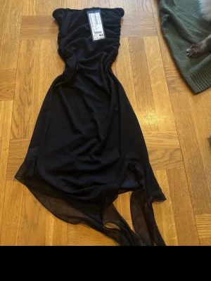 Svart långklänning i chiffong - Elegant svart långklänning med transparenta detaljer nedtill. Klänningen har en mjuk, flytande form och är axelbandslös. Perfekt för dig som vill ha en stilren och tidlös look. Materialet är lätt och ger en snygg siluett.