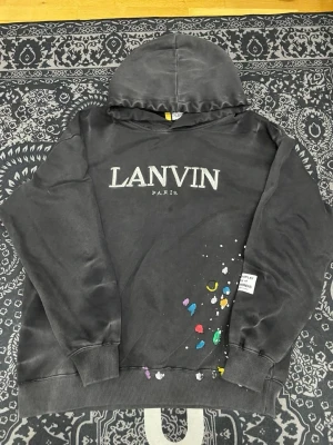 Svart hoodie från Lanvin Paris - Snygg svart hoodie från Lanvin Paris med stor logga framtill och färgglada stänkdetaljer på ena sidan. Hoodien har huva och ribbade muddar vid ärmar och nederkant. Perfekt för dig som gillar streetwear och vill sticka ut med exklusiv design.