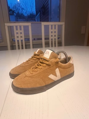 Beige Veja sneakers i mocka - Säljer ett par snygga beige sneakers från Veja i mjuk mocka med vita detaljer och logga på sidan. Skorna har lågt skaft, bruna gummisulor och klassisk snörning. Perfekta för dig som gillar stilrena och trendiga sneakers. Storlek 40 funkar 39,5, har använt några dagar. Kan göra 999kr vid snabbt pris annars är original priset 1299kr. Har inget kvitto eller låda.