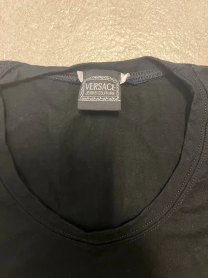 Svart långärmad topp från Versace storlek S - Snygg svart långärmad topp från Versace Jeans Couture med rund halsringning och diskret logga i nacken. Tillverkad i mjuk bomull som känns skön mot huden. Perfekt för dig som gillar stilrena och exklusiva plagg.