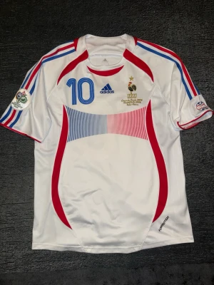 Frankrike tröja 2006 - Säljer en vit matchtröja från Frankrikes landslag, Adidas, med nummer 10 och Zidane på ryggen. Tröjan har blå och röda detaljer, broderade märken och tryck från EM 2005. Korta ärmar och sportig passform, perfekt för fotbollsfans.