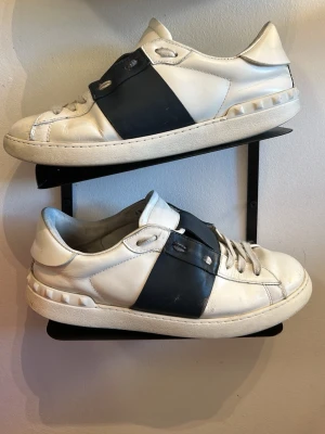 Vit och svart Valentino Garavani sneaker - Snygga sneakers från Valentino Garavani i vitt läder med en bred svart rem över mitten. Klassisk låg modell med rund tå och vita snören. Sulan är vit och har diskreta detaljer längs kanten. Perfekta för dig som gillar stilrena och exklusiva sneakers.