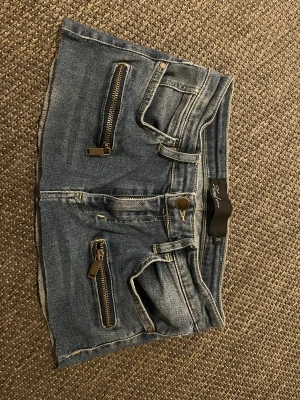 Mini Jeans kjol - Säljer en blå jeanskjol från Zara med två framfickor och dekorativa dragkedjor. Kjolen har klassisk femficksdesign, bälteshällor och rå kant nedtill. Perfekt för dig som gillar en edgy och avslappnad stil.