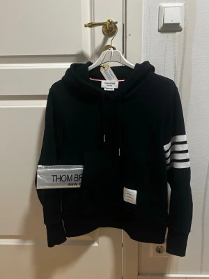 Navy Blue hoodie från Thom Browne - Sitter som XS. Snygg svart hoodie från Thom Browne med klassiska vita ränder på ena ärmen och en stor magficka. Tröjan har huva med dragsko och en vit patch med logga framtill. Perfekt för en clean och stilren look.