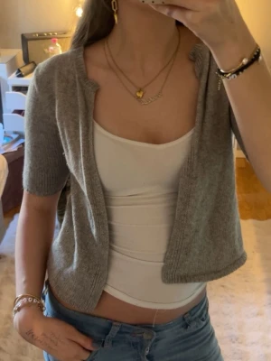 Grå kortärmad stickad kofta - Säljer en grå, kortärmad stickad kofta med öppen front och rund halsringning. Koftan har ribbade kanter och är i mjukt material. 