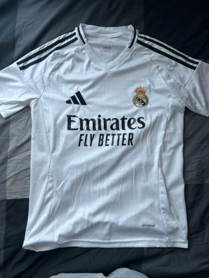 Real Madrid matchtröja Adidas vit - Säljer en vit Real Madrid matchtröja från Adidas med svarta detaljer och klubbmärke på bröstet. Tröjan har svarta ränder på axlarna, rund hals och trycket 'Emirates Fly Better' på framsidan. Tillverkad i lätt och ventilerande material.