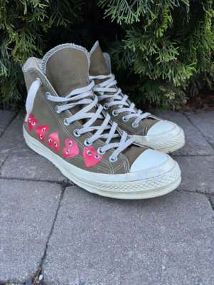 Converse x Comme des Garçons PLAY Chuck 70 High – Khaki/Olive - Säljer nu mina Converse x CDG Play i den snygga och lite mer ovanliga färgen khaki/olivgrön.  Detaljer: • Modell: Chuck Taylor All Star 70 High • Färg: Khaki (Olive) med de klassiska röda hjärtana längs sidan. • Storlek: 42.5 • Nypris: Ca 1 600–1 800 kr. Skick: Skorna är använda men i ett fint begagnat skick. • Sula: Sulan har lite naturligt slitage vid hälarna och sulan (se bild 4), men inget som påverkar användningen eller komforten. Hör av dig vid frågor!