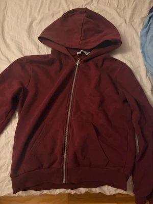 Vinröd hoodie med dragkedja - En enkel och stilren vinröd hoodie med dragkedja framtill och huva. Hoodien har två fickor på magen och ribbade muddar vid ärmslut och nederkant. Perfekt för chill dagar eller när du vill ha något bekvämt.