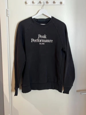 Svart sweatshirt Peak Performance - Klassisk svart sweatshirt från Peak Performance med vit logga och text 'Est. 1986' på bröstet. Tröjan har rund halsringning, ribbade muddar vid ärmslut och nederkant samt en avslappnad passform. Perfekt för en casual och sportig stil.