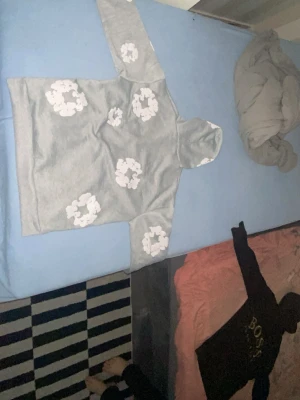 Denim tears grå ( - Säljer en grå hoodie med stora vita blommor tryckta över hela tröjan. Hoodien har en klassisk huva och en magficka framtill. Perfekt för dig som gillar en chill och trendig stil. Materialet känns mjukt och bekvämt, troligen bomullsblandning.  kan vara för bredd om du är för small