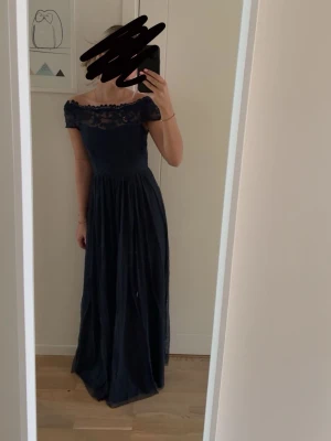 Marinblå långklänning med spets - Elegant marinblå långklänning med spetsdetaljer upptill. Klänningen har en vacker öppen rygg och är figurnära upptill med ett fallande kjolparti. Perfekt för bal eller festliga tillfällen.