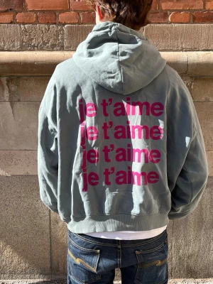 Grå hoodie med rosa text -  oversized grå hoodie med huva. På ryggen står det 'je t’aime' flera gånger i stor rosa text. Hoodien har ribbade muddar och är perfekt för trendig och unik klädstil. 