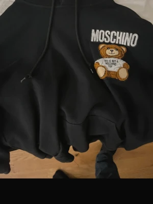 Svart Moschino hoodie med björn - Snygg svart hoodie från Moschino med broderad björn och texten 'This is not a Moschino Toy' på bröstet. Klassisk huva med dragsko och mjukt material som känns riktigt skönt. Perfekt för dig som vill ha en statement hoodie med cool design.