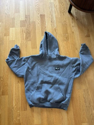 Blå hoodie från PLT med huva - Säljer en blå hoodie från PLT med klassisk huva och stor magficka. Hoodien har en enkel design med PLT-logga på bröstet och ribbade muddar vid ärmslut och nederkant. Perfekt för en avslappnad och chill stil.