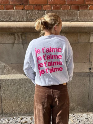 Grå sweatshirt med rosa text - Snygg grå sweatshirt med trycket 'je t’aime' i rosa flera gånger på ryggen. Tröjan har rund halsringning, långa ärmar och ribbade muddar.