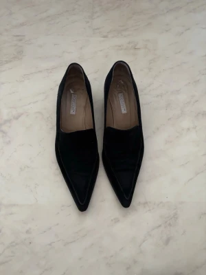 Svarta mockaloafers med klack - Säljer ett par svarta loafers i mjuk mocka. Skorna har spetsig tå och låg klack, vilket ger en stilren och modern look. Insidan är fodrad i beige och de har en klassisk slip-on design. Perfekta för dig som gillar minimalistisk stil.