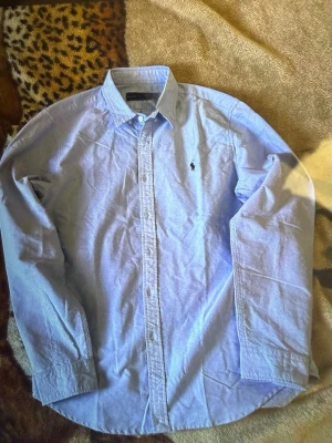 Blå skjorta från Ralph Lauren - Klassisk ljusblå skjorta från Ralph Lauren med button-down krage och ikonisk broderad logga på bröstet. Skjortan har långa ärmar och knäppning framtill. Perfekt till jeans eller chinos för en clean och stilren look.