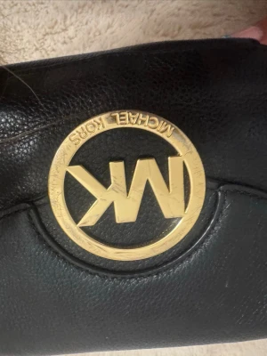 Svart axelväska Michael Kors - Snygg svart axelväska från Michael Kors med stort guldfärgat MK-emblem framtill. Väskan har en kedjedetalj på axelremmen och är gjord i slätt läder. Perfekt för dig som gillar stilrena accessoarer med lyxig känsla.