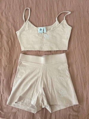 Beige bodyset från Adidas - Säljer ett beige bodyset från Adidas med broderad logga på både topp och shorts. Setet består av en croppad topp med tunna axelband och matchande höga shorts. Materialet är mjukt och stretchigt, perfekt för en avslappnad look.