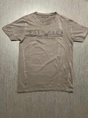 Beige t-shirt Polo Club Saint Vincent - Snygg beige t-shirt från Polo Club Saint Vincent med tryck framtill.Den är bara anständig 1 gång oc den är perfekt till jeans eller shorts för en avslappnad look.