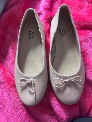 Beige ballerinaskor med rosett - Säljer ett par stilrena beige ballerinaskor med rund tå och söt rosettdetalj framtill. Skorna har en klassisk design och är tillverkade i ett slätt, syntetiskt material. Perfekta för dig som gillar enkel och tidlös stil.