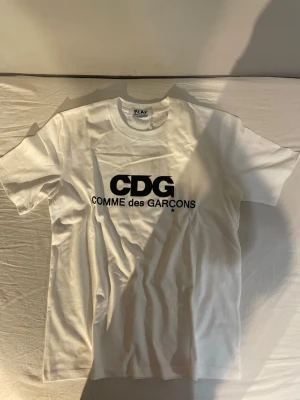 Vit CDG Comme des Garçons t-shirt - Snygg vit t-shirt från Comme des Garçons med stor svart CDG-logga och text på bröstet. Klassisk rund halsringning och korta ärmar. Tillverkad i mjuk bomull för skön känsla och enkel stil.