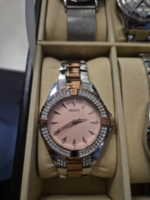 Seksy klocka i roséguld och silver - Snygg klocka från Seksy med roséguldfärgad urtavla och detaljer i både roséguld och silver. Klockan har glittrande stenar runt urtavlan och ett metallarmband som kombinerar två färger för en lyxig look.