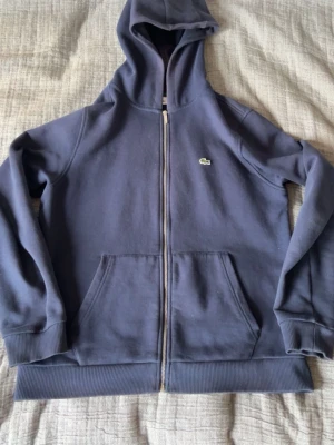 Mörkblå hoodie från Lacoste - Mörkblå hoodie från Lacoste med dragkedja framtill och klassisk krokodil-logga på bröstet. Tröjan har två stora fickor fram, ribbade muddar och en skön huva. Tillverkad i mjukt bomullsmaterial som känns riktigt bekvämt.