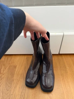 Bruna lackade stövletter med klack - Säljer ett par bruna stövletter i glansigt lackmaterial med fyrkantig tå och chunky klack, tajta runt vristen men i stretchigt material så de går på! Superfina och varsamt använda av mig