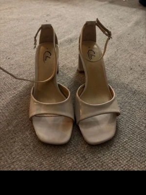 Beige pumps med blockklack från Clou - Snygga beige pumps från Clou med öppen tå, bred rem över foten och justerbart ankelband. Skorna har en stabil blockklack och en stilren, fyrkantig tå. Perfekta för dig som gillar minimalistisk och trendig design.