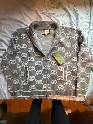 Grå ziptröja - Säljer en grå stickad ziptröja från Gucci med vitt GG-mönster över hela plagget. Tröjan har ribbad krage, muddar och nederkant samt två fickor framtill. Materialet är mjukt och tröjan har en lyxig känsla. Perfekt för dig som gillar exklusiva märken.