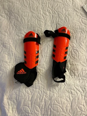 Röda benskydd från Adidas - Säljer ett par röda benskydd från Adidas med svarta detaljer och klassiska ränder. De har justerbara kardborreband och svart vaddering för extra komfort. Perfekta för fotbollsträning eller match.