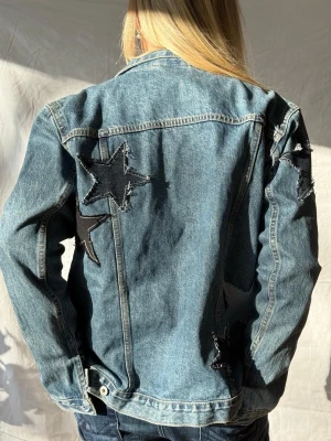 Jeansjacka med stjärnpatchar - Jeansjacka med stora stjärnpatchar i svart och ljusblått på ryggen och ena ärmen. Perfekt för dig som vill sticka ut med en unik detalj på din outfit.🩵
