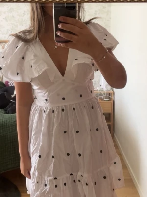 Vit prickig klänning med volang - Superfin vit klänning med svarta prickar och volangdetaljer vid axlarna. Djup v-ringning och kort ärm med puff. Klänningen har en luftig passform och är perfekt för varma dagar. Tillverkad i ett lätt och bekvämt material.