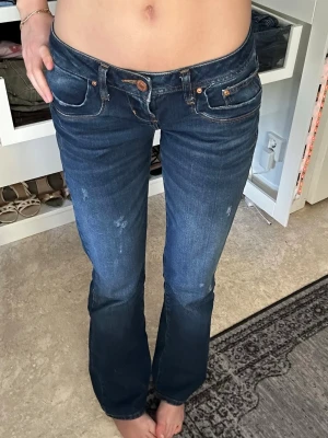 Mörkblå bootcut jeans med låg midja - Säljer ett par mörkblå bootcut jeans med låg midja och slitna detaljer. Jeansen har klassiska fem fickor och kopparfärgade nitar. Perfekta för dig som vill ha ett par jeans som sitter snyggt på höften. Modellen ”valerie” av lbt. 