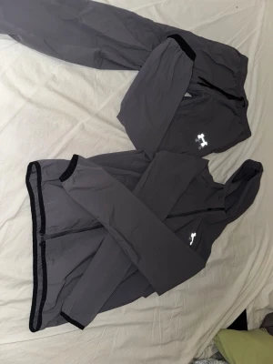 Grå träningsset från Under Armour - Snyggt grått träningsset från Under Armour med matchande jacka och byxor. Jackan har huva, hel dragkedja och svarta detaljer vid ärmslut och nederkant. Byxorna har resår i midjan och logga på benet. Perfekt för träning eller uppvärmning.