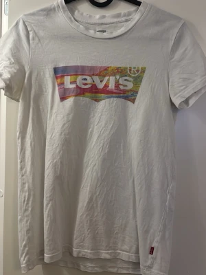 Vit Levi's t-shirt med färgglad logga - Säljer en vit t-shirt från Levi's med en stor, färgglad logga på bröstet i regnbågsfärger. Klassisk rund hals och korta ärmar. Tillverkad i mjuk bomull och har en liten röd Levi's-tag på sidan. Perfekt för en avslappnad och trendig look.
