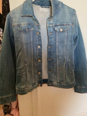 Blå jeansjacka från Here's To The Boys - Klassisk blå jeansjacka från Here's To The Boys med knappar framtill och två bröstfickor med lock. Jackan har långa ärmar och tydliga sömmar som ger en snygg vintagekänsla. Perfekt att slänga över en hoodie eller t-shirt för en chill look.