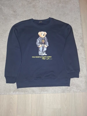Mörkblå sweatshirt Polo Bear Ralph Lauren - Mörkblå sweatshirt från Ralph Lauren med den ikoniska Polo Bear-trycket på bröstet. Tröjan har rund halsringning, ribbade muddar och är tillverkad i mjukt bomullsmaterial. Perfekt för dig som gillar klassisk streetwear med en twist.