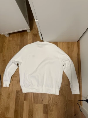 Vit sweatshirt från Nike - Klassisk vit sweatshirt från Nike med rund halsringning och diskret svart Nike-logga på bröstet. Tröjan har långa ärmar, ribbade muddar och är tillverkad i mjukt bomullsmaterial. Perfekt för en clean och sportig look.