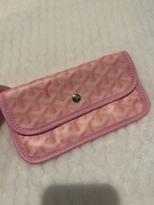 Rosa clutch - Säljer en rosa clutch med det klassiska mönstret och silverfärgad knappstängning. Plånboken har rosa kanter och insidan är ljus med plats för kort och sedlar. Materialet är canvas med läderdetaljer. Perfekt för dig som gillar lyxiga accessoarer.