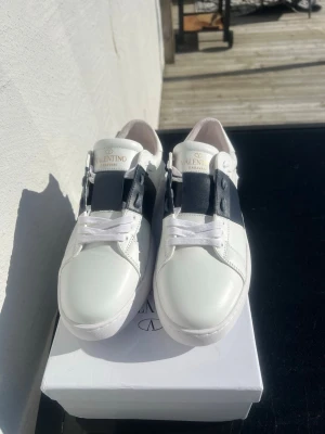 Valentino vit och blåa sneakers  - Snygga vita sneakers från Valentino med ett brett svart band över mitten och coola nitar på hälen. Skorna är i läder och har klassisk låg profil med vita snören och diskret logga på plösen. Perfekta för dig som vill ha en clean men ändå unik look.