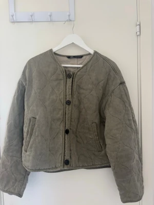 Olivgrön quiltad cardigan från Zara - Quiltad cardiganjacka från Zara i olivgrönt tyg med rund halsringning och stora svarta knappar framtill. Jackan har två fickor och är fodrad på insidan. Perfekt för lager på lager och ger en avslappnad men trendig look.