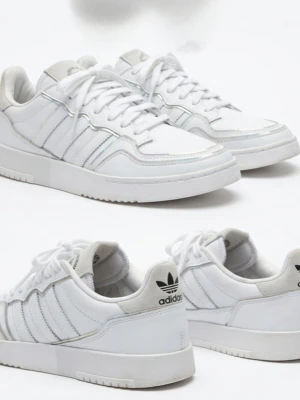 Vita Adidas sneakers med holografiska detaljer - Säljer ett par vita Adidas sneakers i lågt snitt med klassiska tre ränder på sidan och holografiska detaljer längs sömmarna. Skorna har en chunky sula, snörning och Adidas-logga på hälen. Tillverkade i läder och textil för en clean och modern look.