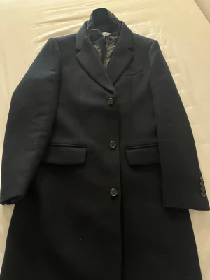 Svart rock från Selected Homme - Stilren svart rock från Selected Homme med tre knappar framtill, klassisk krage och två fickor med lock. Rocken har en avtagbar innerkrage i quiltat material för extra värme och stil. Perfekt för dig som vill ha en tidlös och snygg look. Endast använd vid ett tillfälle