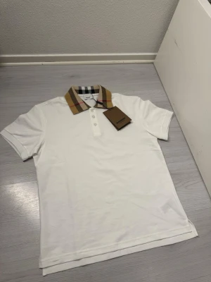 Vit Burberry pikétröja med rutig krage - Säljer en vit pikétröja från Burberry med den klassiska beige, svarta och röda rutiga kragen. Tröjan har korta ärmar, knappar framtill och är tillverkad i mjuk bomull. Perfekt för dig som vill ha en stilren och ikonisk look. Längden är 64 cm bredden fårn axel till axel är 51 cm. Kan sänkas vid snabb affär 